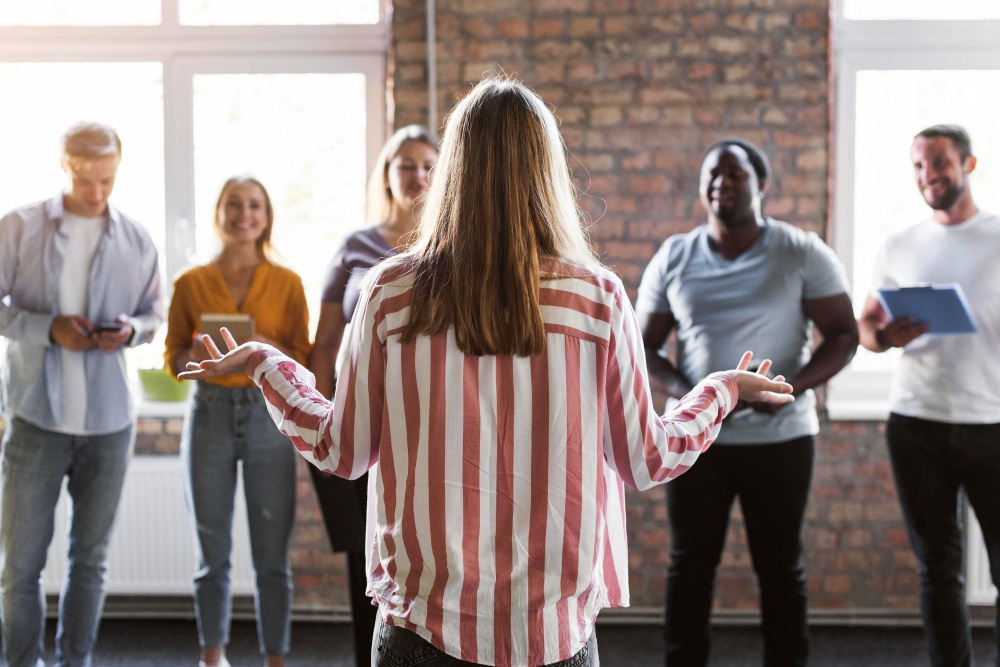 Personne de dos en chemise rayée rouge et blanche s'exprimant devant un groupe de collègues souriants, dans un environnement de travail moderne avec mur en briques apparentes.